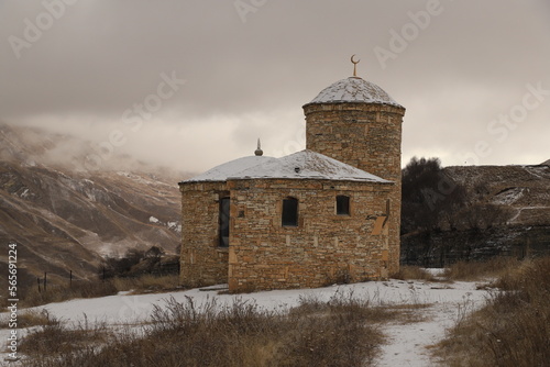 An ancient stone Muslim temple in the mountains of Dagestan - stands on a snowy slope against the backdrop of mountains and a gloomy sky.