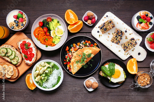 Healthy breakfast food table scene. Overhead view on a dark wood background. Omelette, nutritious bowl, toasts, granola bars, smoothie bowl, yogurts and fruits.
