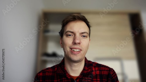 A young man in a red plaid shirt is sitting in front of a laptop webcam. The background is a mess, dirt. The guy gets upset, turns on the digital background blur, looks at the result with smile.