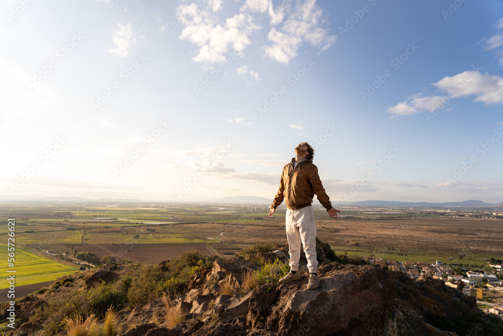 Fototapeta premium Young man on the top of a mountain taking a deep breath with his head high and arms open feeling the sun, air and wind on his body. Concept of freedom. Copy space on the left.