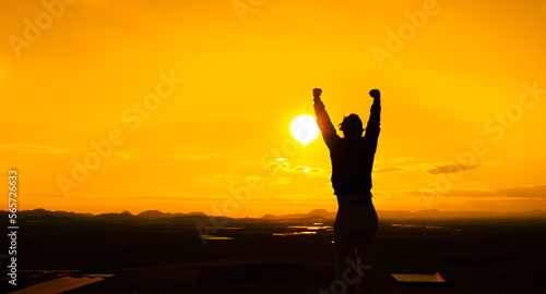 Person with arms up as a sign of self-improvement and motivation. Silhouette of a man at sunset on top of a mountain. Warm orange sky. Concept purposes, challenges. Copy space to the left