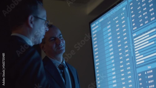 Cheerful businessmen analyzing big data on big digital screen. Financial analysts talking in modern monitoring room, solving work issues, processing information in office. Occupation concept 