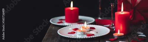 Saint Valentine's Day celebration. Red burning candles, hearts, gift box, postcard on dark wooden background. Happy holiday. Table decor for festive dinner, romantic atmosphere. Banner photo