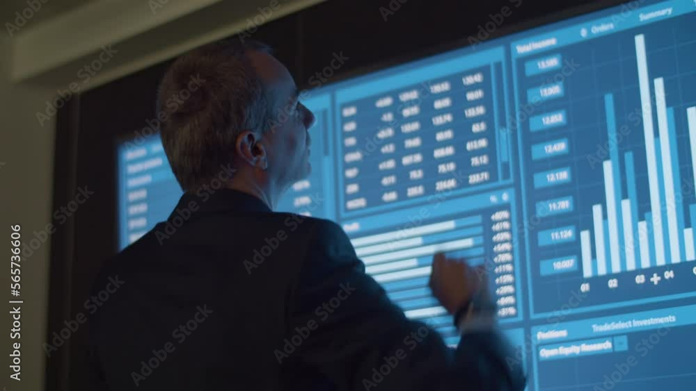 Businessman analyzing data and statistics shown on big screen. Back view of male manager in official suit in dark monitoring room working on project and processing information. Technology concept Businessman analyzing data and statistics shown on big screen. Back view of male manager in official suit in dark monitoring room working on project and processing information. Technology concept