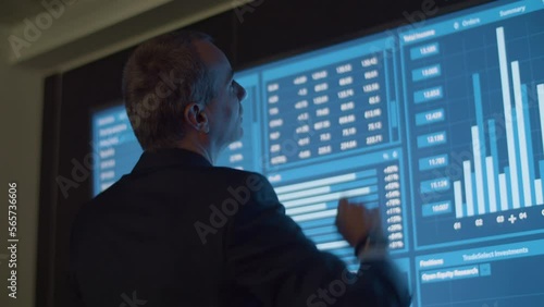 Businessman analyzing data and statistics shown on big screen. Back view of male manager in official suit in dark monitoring room working on project and processing information. Technology concept