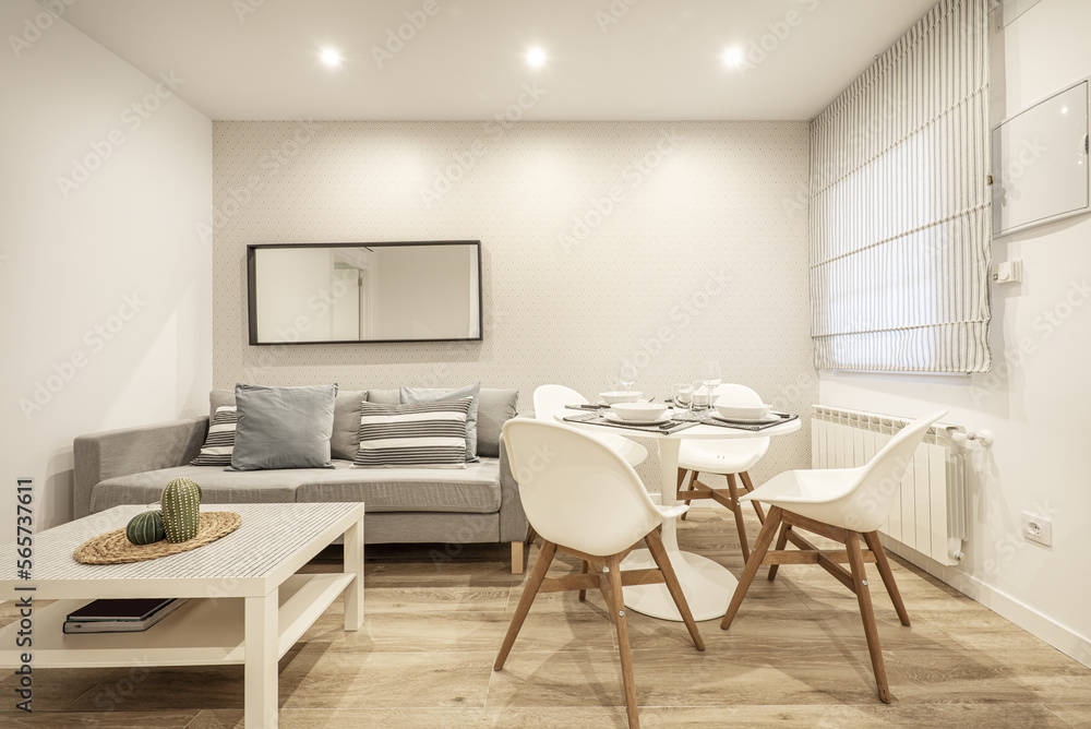 Living-dining room of a house furnished with a gray sofa, a window with a fabric blind and a round dining table Living-dining room of a house furnished with a gray sofa, a window with a fabric blind and a round dining table