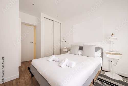 Bedroom with a gray fabric double bed and white duvet with clean rolled towels and a built-in wardrobe with white wooden sliding doors