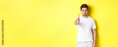 Confident man saying no, decline and prohibit something, showing one finger and frowning, standing over yellow background