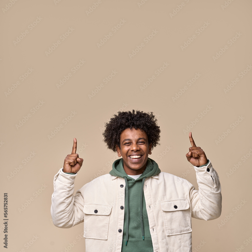 Smiling cool ethnic guy pointing fingers up isolated on beige background. Happy African American teenager fashion model, student, hipster presenting marketing offer, advertising shopping sale.