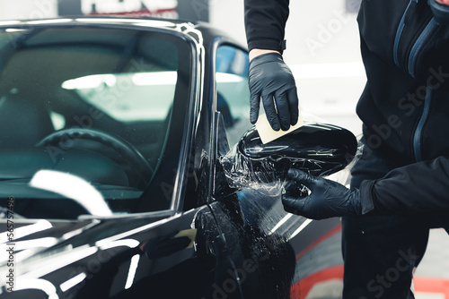 Man wearing black gloves leaning over to smooth out PPF Paint Protection Film applied to side mirror of black car. Professional car detailing. Horizontal indoor shot. High quality photo