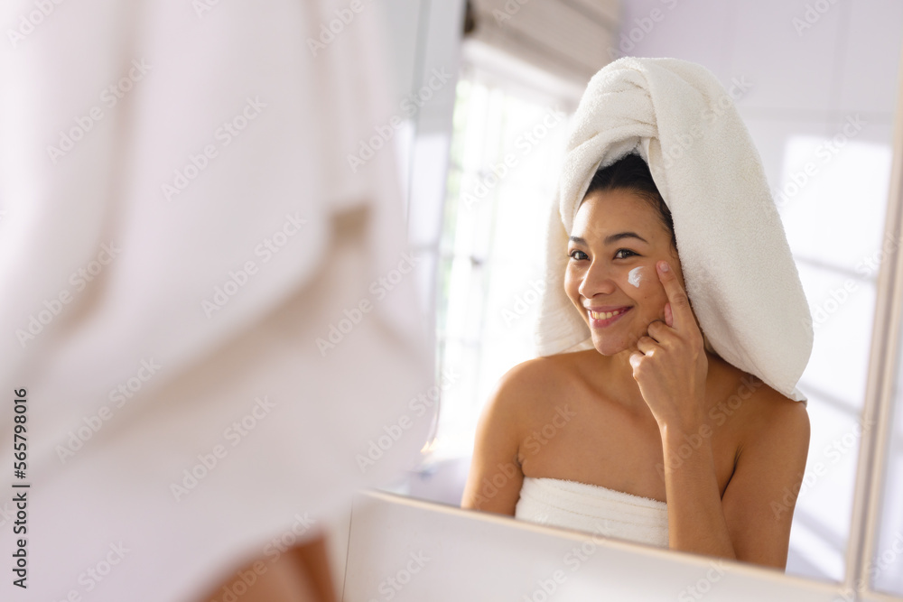 Happy biracial woman wearing towel smiling in bathroom mirror moisturising face, with copy space Happy biracial woman wearing towel smiling in bathroom mirror moisturising face, with copy space
