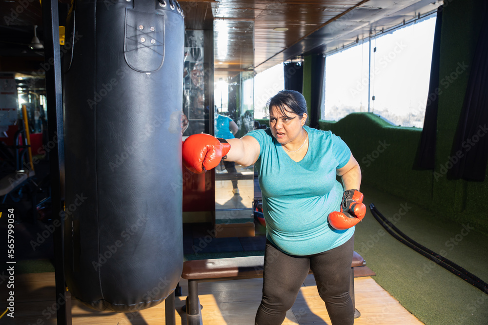 Young overweight indian woman wearing boxing gloves punching bag in gym, Plus size sporty asian female workout to lose weight. Fitness and healthcare.