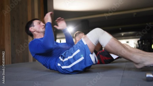 Athletic men at gym doing a series of active sit-ups. Fit male fighters in sportswear performing exhausting workout session against the wall. Sportsmen doing sit-ps and crunches photo