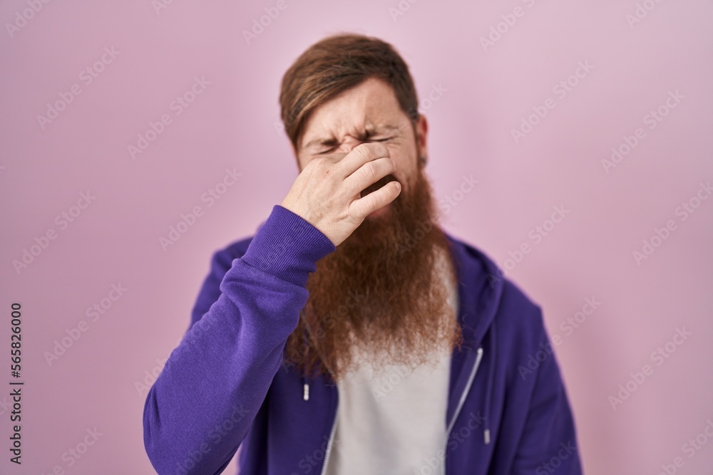 Caucasian man with long beard standing over pink background smelling something stinky and disgusting, intolerable smell, holding breath with fingers on nose. bad smell Caucasian man with long beard standing over pink background smelling something stinky and disgusting, intolerable smell, holding breath with fingers on nose. bad smell