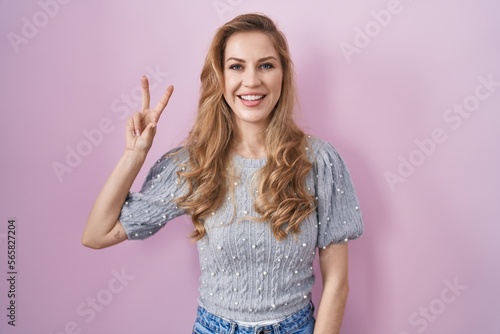Beautiful blonde woman standing over pink background showing and pointing up with fingers number two while smiling confident and happy.