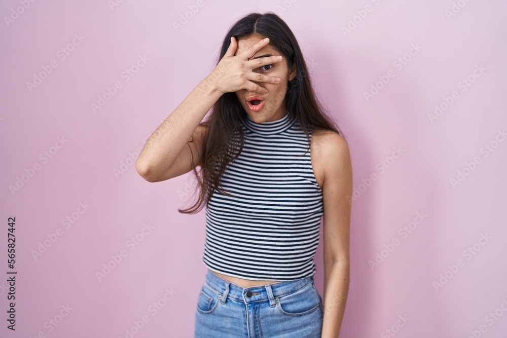 Young teenager girl wearing casual striped t shirt peeking in shock covering face and eyes with hand, looking through fingers with embarrassed expression. Young teenager girl wearing casual striped t shirt peeking in shock covering face and eyes with hand, looking through fingers with embarrassed expression.