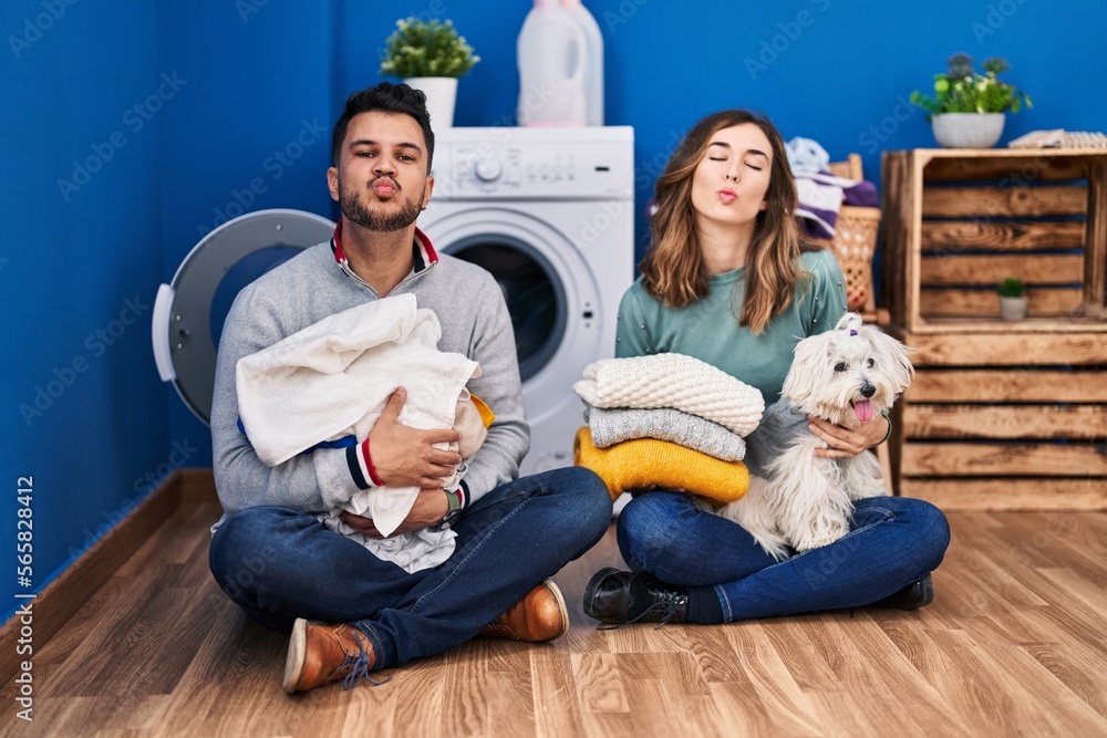 Young hispanic couple doing laundry sitting on the floor looking at the camera blowing a kiss being lovely and sexy. love expression. Young hispanic couple doing laundry sitting on the floor looking at the camera blowing a kiss being lovely and sexy. love expression.
