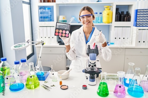 Young blonde woman working at scientist laboratory with make up smiling happy and positive  thumb up doing excellent and approval sign