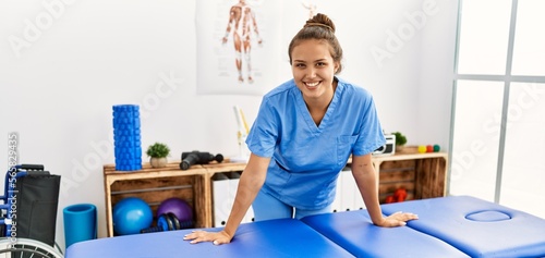 Young beautiful hispanic woman physiotherapist smiling confident standing at rehab clinic photo