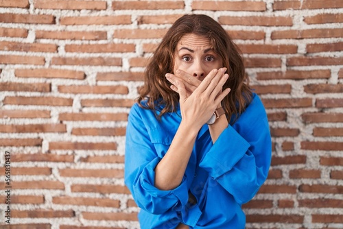 Beautiful brunette woman standing over bricks wall shocked covering mouth with hands for mistake. secret concept.