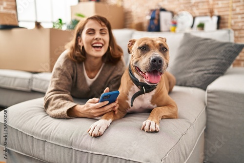 Young caucasian woman using smartphone lying on sofa with dog at new home
