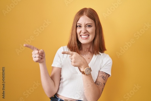 Young redhead woman standing over yellow background pointing aside worried and nervous with both hands, concerned and surprised expression