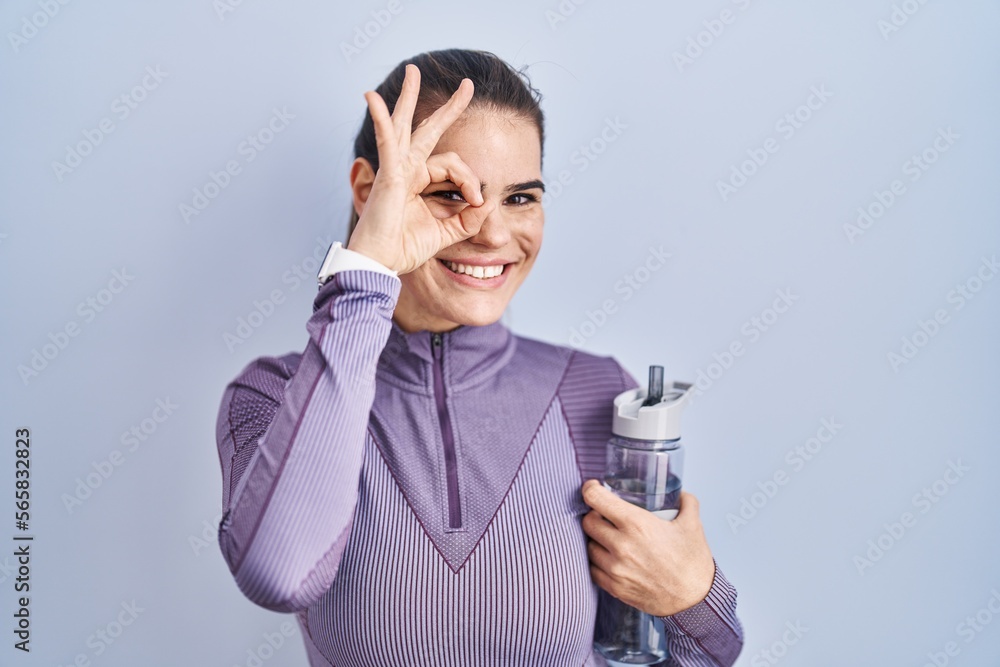 Beautiful woman wearing sportswear holding water bottle doing ok gesture with hand smiling, eye looking through fingers with happy face.