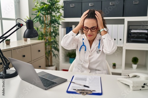 Young hispanic woman wearing doctor uniform and stethoscope suffering from headache desperate and stressed because pain and migraine. hands on head.