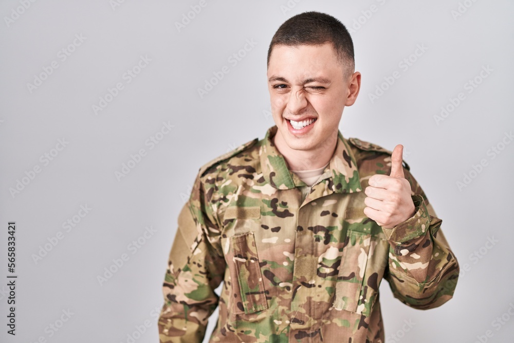 Young man wearing camouflage army uniform doing happy thumbs up gesture with hand. approving expression looking at the camera showing success.