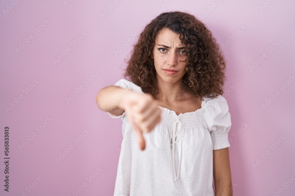 Hispanic woman with curly hair standing over pink background looking unhappy and angry showing rejection and negative with thumbs down gesture. bad expression. Hispanic woman with curly hair standing over pink background looking unhappy and angry showing rejection and negative with thumbs down gesture. bad expression.