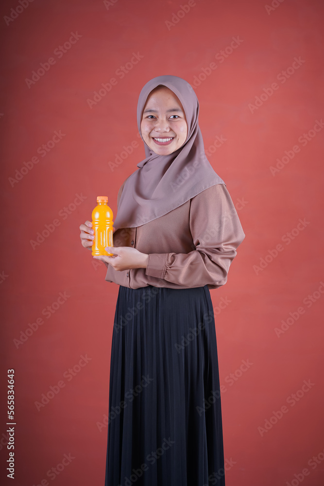 Beautiful Asian woman in brown shirt and hijab smiling cheerfully holding bottle of orange juice on brown background.