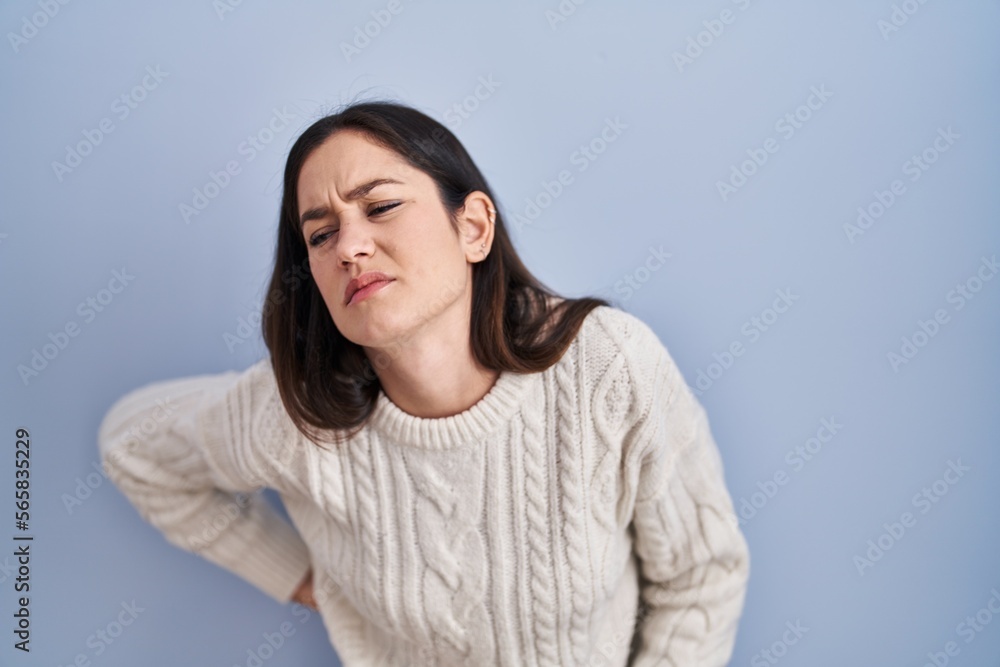 Young brunette woman standing over blue background suffering of backache, touching back with hand, muscular pain Young brunette woman standing over blue background suffering of backache, touching back with hand, muscular pain