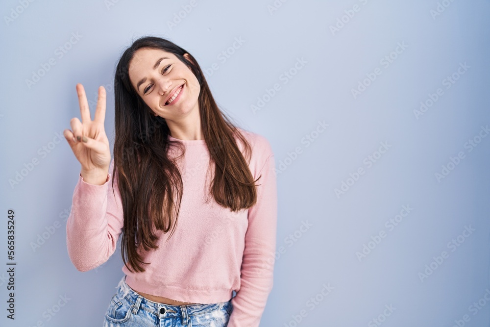 Young brunette woman standing over blue background smiling looking to the camera showing fingers doing victory sign. number two.