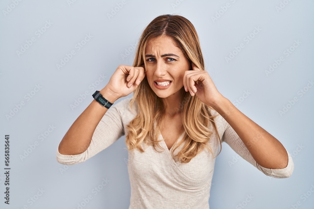 Young blonde woman standing over isolated background covering ears with fingers with annoyed expression for the noise of loud music. deaf concept. Young blonde woman standing over isolated background covering ears with fingers with annoyed expression for the noise of loud music. deaf concept.