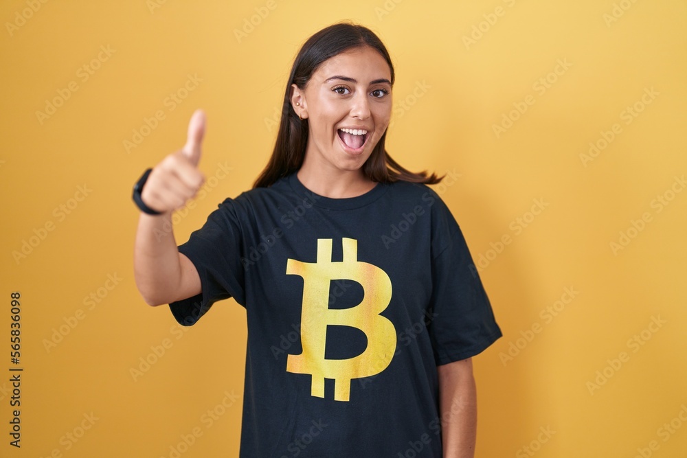 Young hispanic woman wearing bitcoin t shirt approving doing positive gesture with hand, thumbs up smiling and happy for success. winner gesture. Young hispanic woman wearing bitcoin t shirt approving doing positive gesture with hand, thumbs up smiling and happy for success. winner gesture.