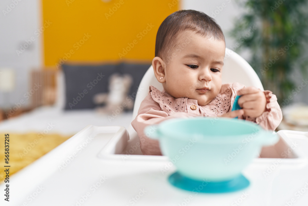 Adorable hispanic baby sitting on highchair holding spoon at bedroom Adorable hispanic baby sitting on highchair holding spoon at bedroom