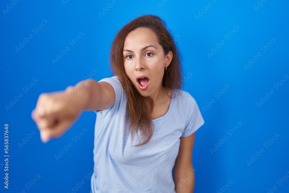 Brunette woman standing over blue background pointing with finger surprised ahead, open mouth amazed expression, something on the front Brunette woman standing over blue background pointing with finger surprised ahead, open mouth amazed expression, something on the front