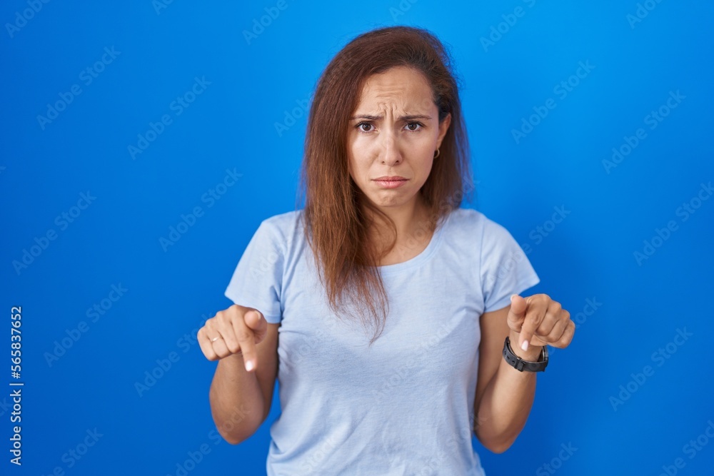 Brunette woman standing over blue background pointing down looking sad and upset, indicating direction with fingers, unhappy and depressed. Brunette woman standing over blue background pointing down looking sad and upset, indicating direction with fingers, unhappy and depressed.