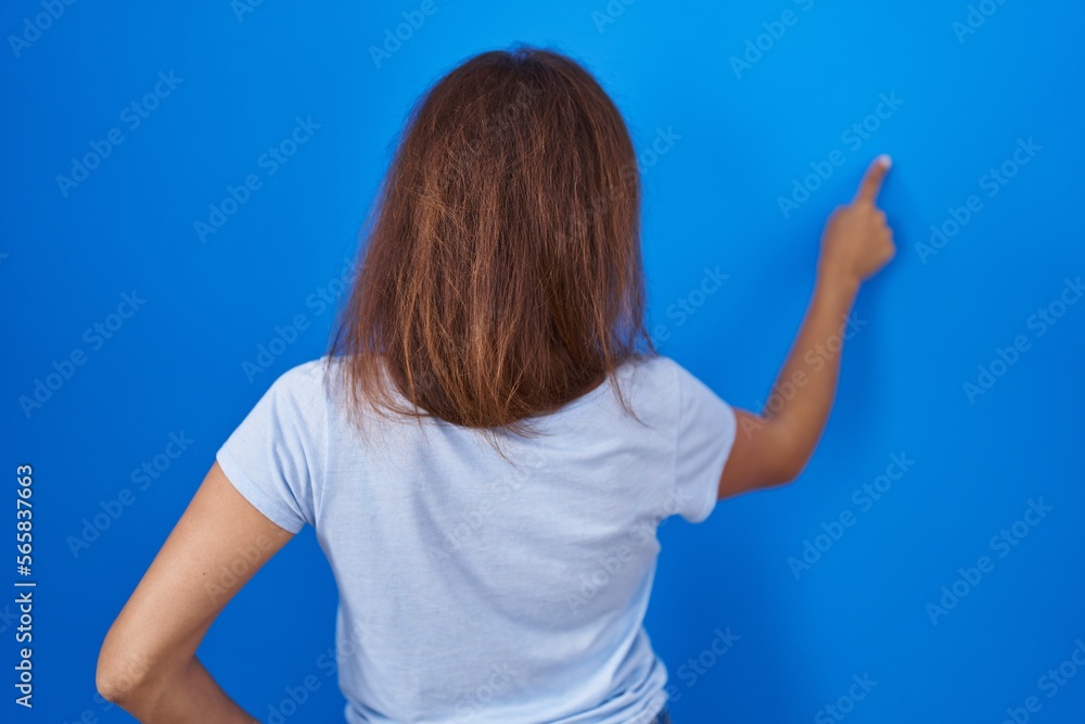 Fototapeta premium Brunette woman standing over blue background posing backwards pointing ahead with finger hand