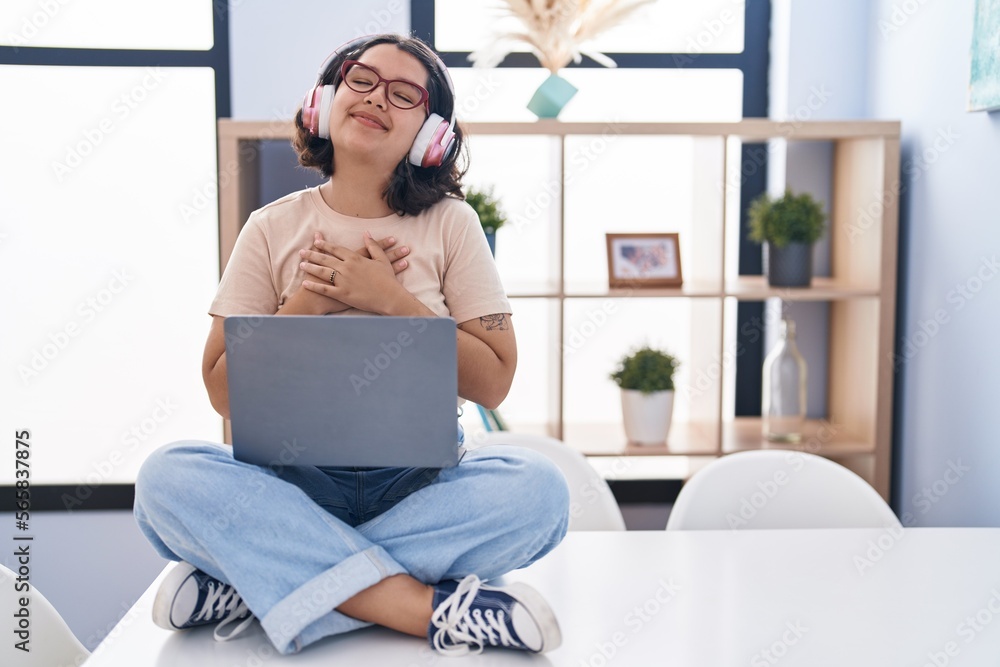 Young hispanic woman using laptop sitting on the table wearing headphones smiling with hands on chest with closed eyes and grateful gesture on face. health concept. Young hispanic woman using laptop sitting on the table wearing headphones smiling with hands on chest with closed eyes and grateful gesture on face. health concept.