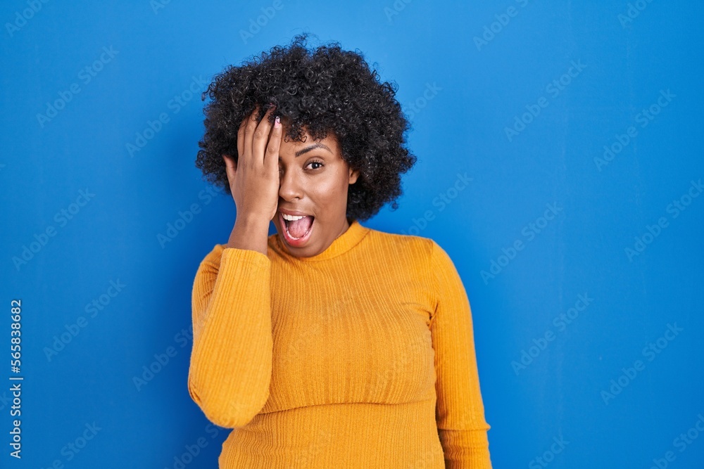 Black woman with curly hair standing over blue background covering one eye with hand, confident smile on face and surprise emotion. Black woman with curly hair standing over blue background covering one eye with hand, confident smile on face and surprise emotion.