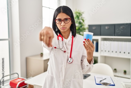 Young hispanic doctor woman holding cotton buds pointing with finger to the camera and to you, confident gesture looking serious