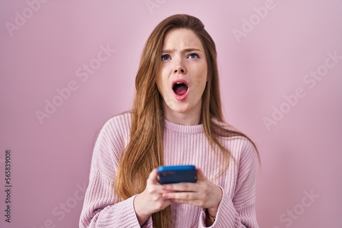 Young caucasian woman using smartphone typing message angry and mad screaming frustrated and furious, shouting with anger looking up.