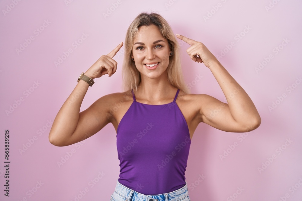 Young blonde woman standing over pink background smiling pointing to head with both hands finger, great idea or thought, good memory Young blonde woman standing over pink background smiling pointing to head with both hands finger, great idea or thought, good memory