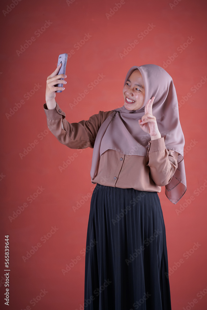 Beautiful Asian woman in brown shirt and hijab smiling cheerfully taking selfie using smartphone against brown background Beautiful Asian woman in brown shirt and hijab smiling cheerfully taking selfie using smartphone against brown background