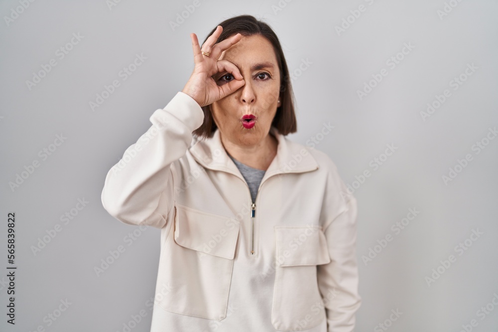 Middle age hispanic woman standing over isolated background doing ok gesture shocked with surprised face, eye looking through fingers. unbelieving expression.