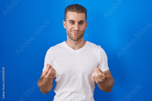 Young caucasian man standing over blue background doing money gesture with hands, asking for salary payment, millionaire business