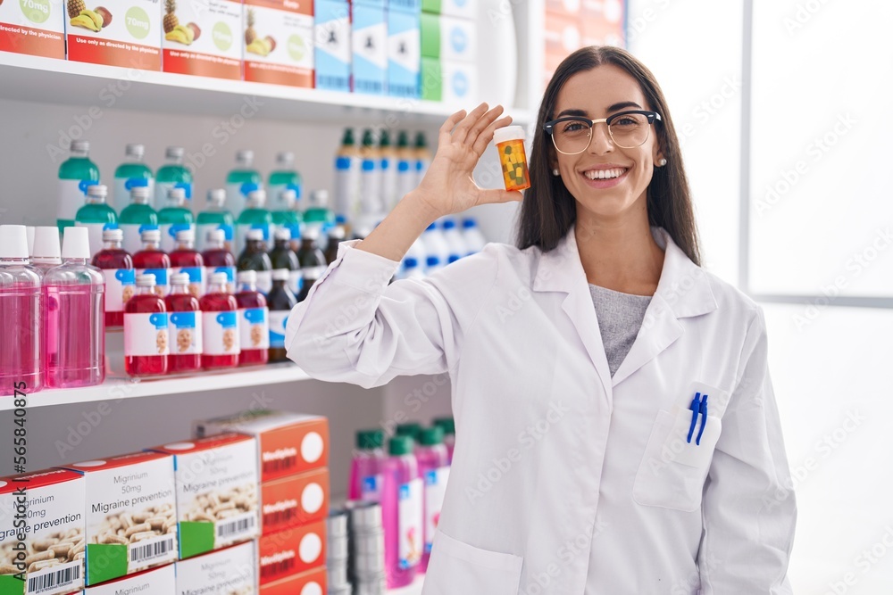 Young beautiful hispanic woman pharmacist smiling confident holding pills bottle at pharmacy