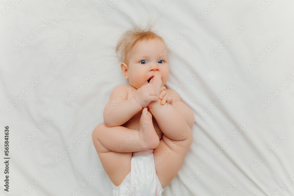 a happy joyful baby in diapers is lying on a white bed and holding his leg. High-quality photo. place for text a happy joyful baby in diapers is lying on a white bed and holding his leg. High-quality photo. place for text