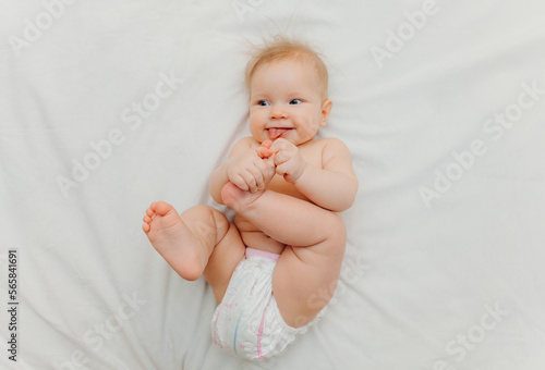 a happy joyful baby in diapers is lying on a white bed and holding his leg. High-quality photo. place for text